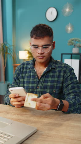 Smiling-Indian-man-using-bank-credit-card-and-smartphone-for-online-shopping-payments-at-home-office