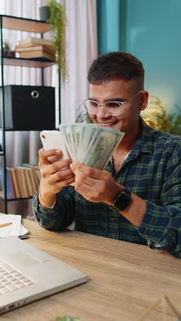Un-Hombre-Feliz-Y-Sonriente-Contando-Dinero-En-Efectivo-En-Dólares-Usando-Ingresos-Por-Teléfono-Inteligente-Ahorra-La-Lotería-En-Casa