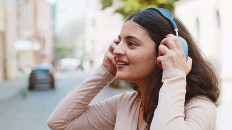 Happy-Indian-young-woman-wearing-wireless-headphones-listening-music-dancing-on-city-street-outdoors