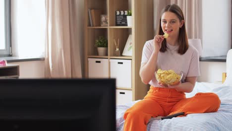 Happy-Girl-with-Crisps-Watching-Tv-at-Home.unhealthy-eating,-fast-food-and-people-concept-happy-girl-with-remote-control-and-crisps-watching-tv-sitting-on-bed-at-home