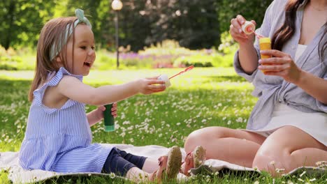 Mother-with-Daughter-Blowing-Soap-Bubbles-at-Park.family,-motherhood-and-people-concept-happy-mother-with-little-daughter-blowing-soap-bubbles-at-summer-park-or-garden