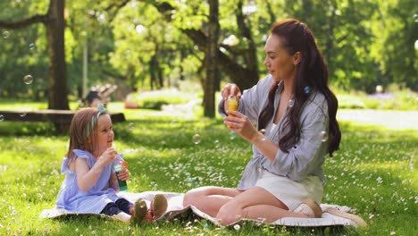 Mother-with-Daughter-Blowing-Soap-Bubbles-at-Park.family,-motherhood-and-people-concept-happy-mother-with-little-daughter-blowing-soap-bubbles-at-summer-park-or-garden