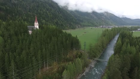 Eine-Drohnenaufnahme-Einer-Kirche-In-Der-Nähe-Eines-Flusses-An-Den-Alpen