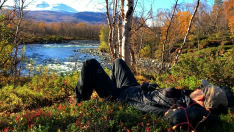 Hiker-taking-a-break-lying-down-by-a-river,-watching-the-scenic-landscape-in-Abisko,-Lapland-in-northern-Sweden