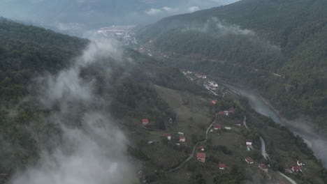 Fly-over-clouds-river-stream-houses-and-lush-dark-forest-tree-in-the-balkans