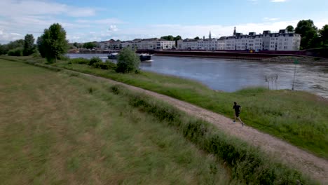 Male-trail-runner-taking-off-along-river-IJssel-seen-from-above-in-Dutch-flatland-with-countenance-of-tower-town-Zutphen-behind