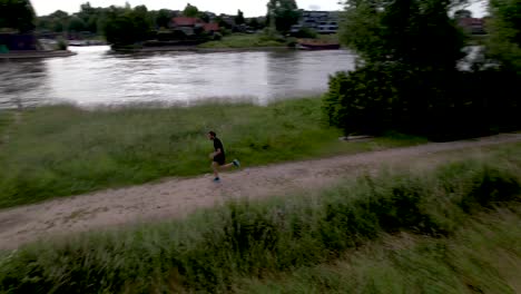 Aerial-following-of-male-trail-runner-taking-off-along-river-IJssel-in-Dutch-flatland-with-countenance-of-tower-town-Zutphen-behind