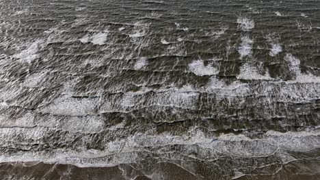 Wide-aerial-tilt-down-view-of-waves-crashing-on-the-beach-with-foam-and-sets-of-surf-rolling-in-at-sunset