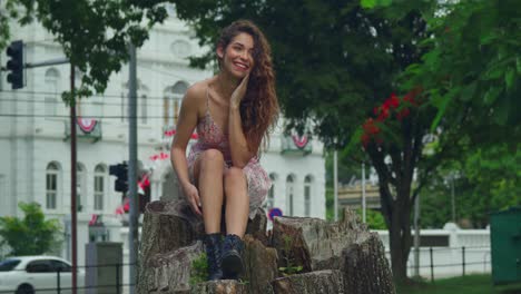 Pretty-curly-hair-latina-sits-on-a-tree-stump-and-smiles-in-a-mini-dress-with-a-castle-in-the-background-at-a-park