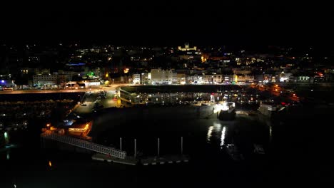 Vuelo-Nocturno-Al-Puerto-De-St-Peter,-Puerto-De-Guernsey,-Con-El-Puerto-Deportivo-De-Los-Visitantes-Y-El-Paseo-Marítimo-Con-Edificios-Iluminados-En-El-Fondo,-Incluido-El-Colegio-Elizabeth