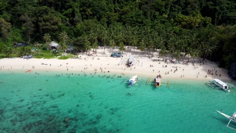 Luftaufnahme:-Philippinische-Banca-Boote-Im-Katamaran-Stil-Kreuzen-Im-Vordergrund-Des-Tropical-Seven-Commando-Beach,-Touristen-Schwimmen-In-Türkisfarbenem,-Klarem-Wasser-Auf-Weißem-Sand