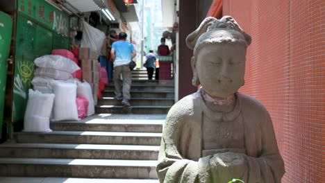 Pedestrians-walk-up-the-stairs-on-a-narrow-street-as-they-pass-a-stone-Buddha-statue-in-a-district-known-for-its-numerous-antique-shops