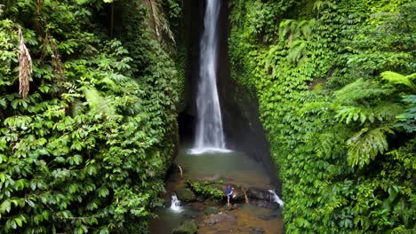 Vista-Aérea-Cinematográfica:-Joven-Caminando-Hacia-Una-Roca-En-El-Fondo-De-La-Cascada-Leke-Leke-En-La-Regencia-De-Tabanan-En-Bali,-Indonesia