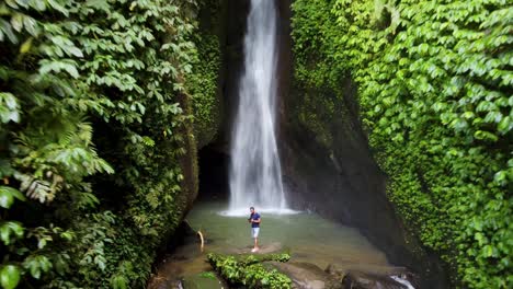 4k-Aéreo:-Joven-Turista-Parado-En-La-Cascada-De-Leke-Leke,-Paraíso-Tropical-Escondido-En-Un-Exuberante-Cañón-Cubierto-De-Musgo-Verde-En-Las-Selvas-De-Bali