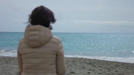 Rear-view-of-woman-wearing-beige-winter-jacket-looking-pensive-at-sea-at-beach