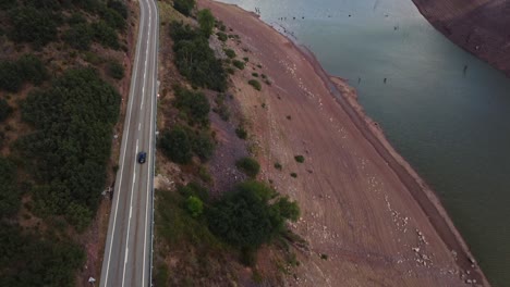 Spectacular-drone-video-of-a-car-traversing-scenic-roads-by-a-lake,-showcasing-an-impressive-hanging-bridge