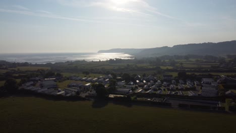 Aerial-view-rising-over-Welsh-caravan-park-at-sunrise-overlooking-shimmering-coastal-bay-and-rolling-countryside-hills