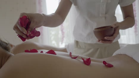 Professional-rose-petals-aromatic-spa-massage,-therapist-is-putting-flower-parts-on-the-back-of-the-laying-person-on-the-massage-table,-well-being-and-health-concept
