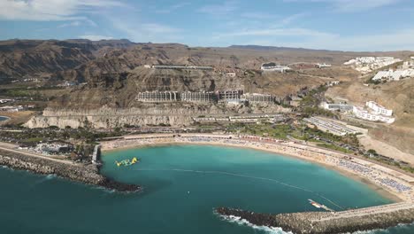 Aerial-backwards-shot-of-beautiful-bay-with-sandy-beach-and-resort-on-hill-during-sunny-day---Playa-de-Amadores,-Gran-Canaria