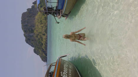Athletic-blonde-woman-walks-into-clear-ocean-between-traditional-Thai-boats