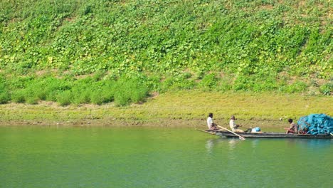 Bengali-Fishermen-and-Villagers-Rowing-a-Long-Pirogue-along-a-Slowly-Flowing-River-in-front-of-a-Vividly-Vegetalised-Riverbank,-Bangladesh