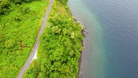 Aerial-drone-of-traffic-of-car-and-motorbikes-driving-along-beautiful-coastal-road-through-dense-green-trees-on-tropical-island-of-Alor-Island,-East-Nusa-Tenggara,-Indonesia