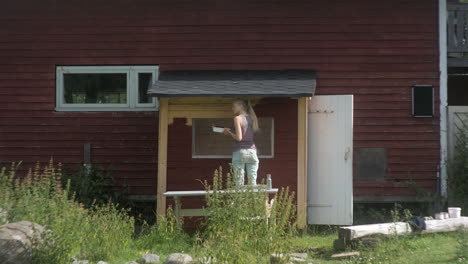 Wide-Shot-of-Beautiful-Blonde-Girl-Painting-a-Red-Traditional-Finish-House,-Nordic-Woman-Doing-Maintenance-Work,-Finland,-Scandinavia