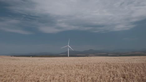 A-wind-turbine-rotating-on-a-wheat-field-with-blue-skies-clouds-and-mountains