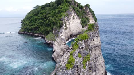 Waves-breaking-on-cliffs-of-lush-green-limestone-sea-mount-rock-formation-against-Atuh-Beach,-Nusa-Penida-Island