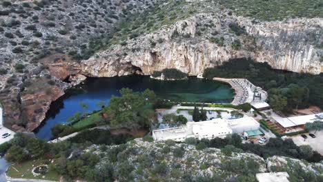 High-angle-bird’s-eye-view-of-the-submerged-lake-of-Vouliagmeni-in-Athens,-Greece-with-stunning-surrounding-cliffs-and-a-highway-above-the-lake-|-4K