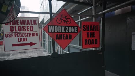 In-a-construction-scaffold-walkway,-signs-warn-of-a-closed-sidewalk-and-upcoming-work-zone,-urging-shared-road-usage