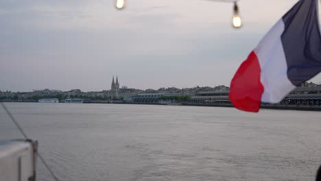 French-flag-waving-on-the-back-of-a-barge-boat-on-the-Garonne-River-in-Bordeaux-France-at-dusk,-Wide-shot