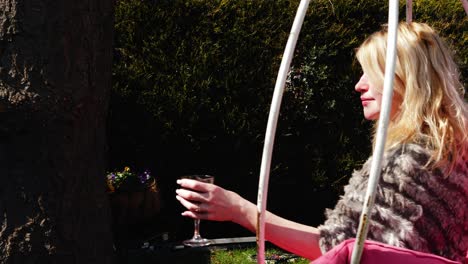 Attractive-woman-enjoys-glass-of-wine-in-garden-chair-swing-wide-4k-slow-motion