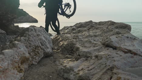 man-carefully-carries-his-bike-over-rocks,-showcasing-his-determination-and-resilience-near-sea-coast
