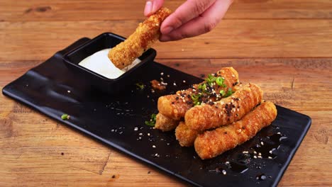 Close-up-of-a-plate-of-cheese-fingers-breaded-with-sesame-seeds,-habanero-peppers,-ranch-dressing-and-Worcestershire-sauce,-a-hand-takes-a-cheese-finger-and-dips-it-in-the-dressing