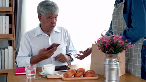 Hombre-Asiático-Senior-Activo-Que-Trabaja-En-Una-Cafetería-Usando-Un-Dispositivo-Móvil,-Reloj-Inteligente-Para-Nfc,-Escaneo-De-Código-Qr,-Pago-Sin-Contacto-Para-Pagar-Café-Y-Bocadillos
