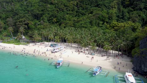 Aerial-view:-catamaran-style-Filipino-banca-boats-traversing-in-foreground-of-Tropical-Seven-Commando-beach-,-Tourists-swimming-in-turquoise-clear-water-on-white-sand