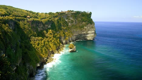 Reveal-shot-of-the-cliffs-at-Uluwatu,-near-the-Uluwatu-temple-with-brilliant-green-waves-crashing-against-the-greenery-covered-cliff-walls