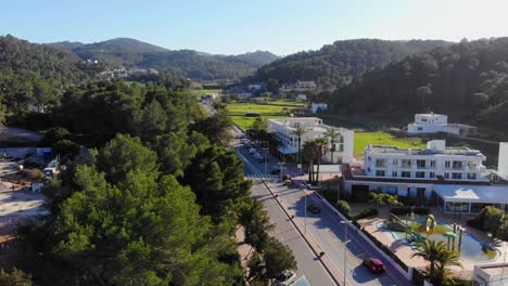 Drone-Descendiendo-Por-Una-Carretera-Con-Algunas-Casas-Con-Una-Piscina-Verde-En-Ibiza