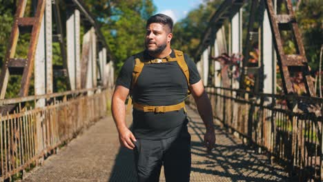Front-view-young-man-traveler-with-backpack-walking-on-rusty-old-bridge-outdoors-on-sunny-summer-day