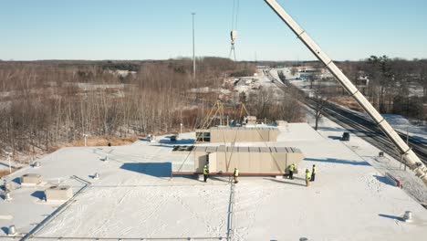 Workers-installing-a-HVAC-unit-tied-to-a-crane-on-an-industrial-building-roof,-aerial