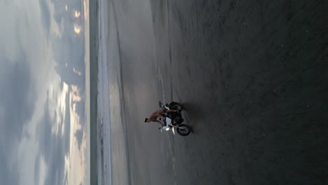 Sweeping-vertical-format-aerial:-Motorcyclists-rides-on-beach-sand