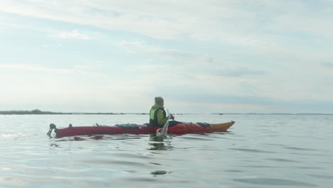 Young-Blonde-Woman-Paddling-in-a-Red-Sea-Kayak-on-the-open-Sea-in-Finland,-Vaasa,-Archipelago,-Beautiful-Summer-Sunset-Atmosphere,-Wide-Shot