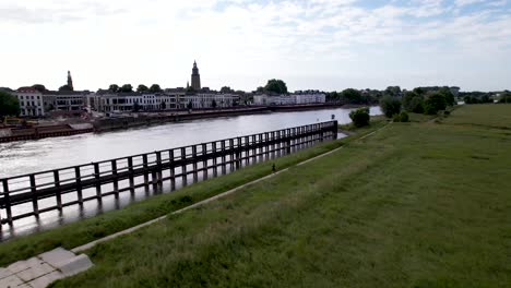 Vast-Dutch-landscape-aerial-following-male-trail-runner-along-river-IJssel-in-floodlands-with-countenance-of-tower-town-Zutphen-behind