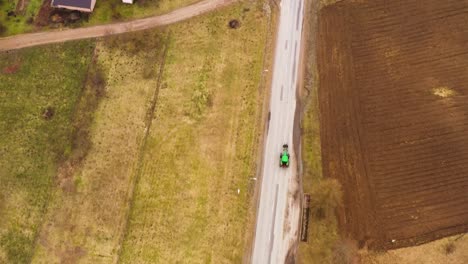 Green-tractor-driving-on-a-countryside-road-passing-houses,-drone-view