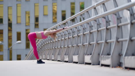 Young-blonde-woman-in-pink-sportswear-does-stretches-on-bridge,-wide