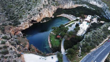 Pan-Bird’s-eye-view-of-the-submerged-lake-Vouliagmeni-in-Athens,-Greece-with-stunning-surrounding-cliffs-and-the-highway-above-the-lake-|-4K