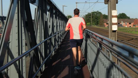 Following-middle-aged-male-trail-runner-on-steel-bridge-between-train-tracks-and-asphalt-road
