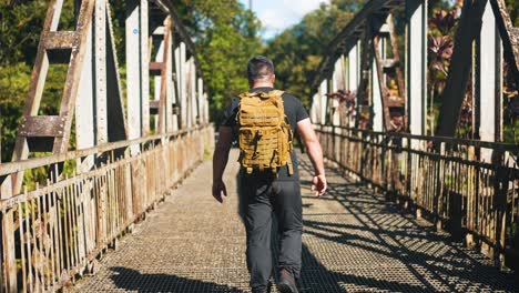 Following-young-man-with-backpack-walking-on-rusty-old-bridge-outdoors-on-sunny-summer-day
