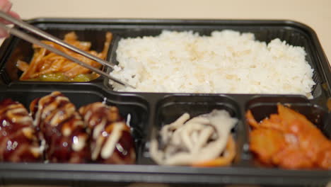 Chopsticks-picking-up-rice-from-Dosirak-or-Korean-lunchbox-with-cutlets-and-kimchi---close-up
